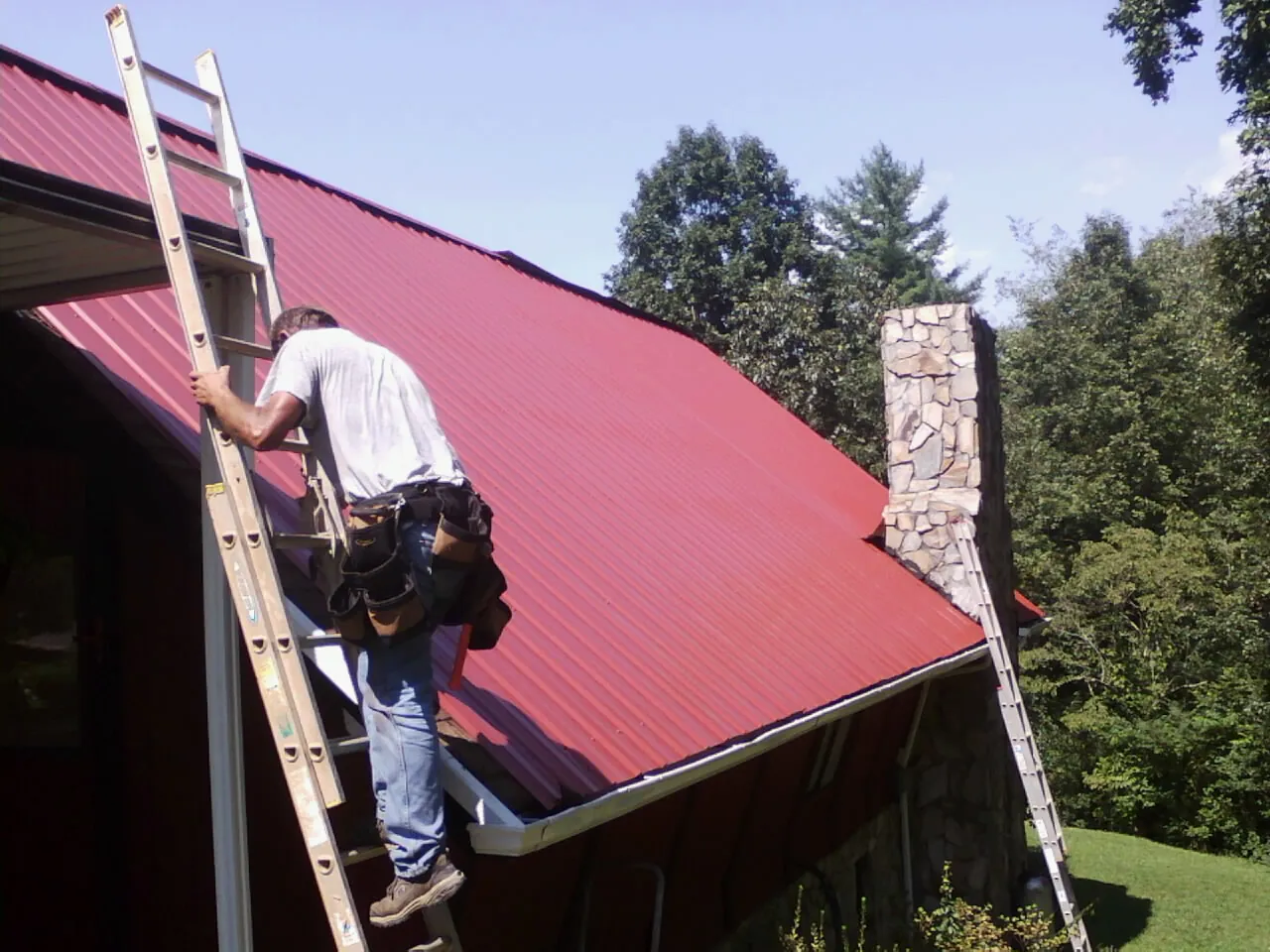Roofer on a ladder beside a red metal roof and stone chimney — Soffit & Fascia Repair by Roofing Brothers in Garrett Park